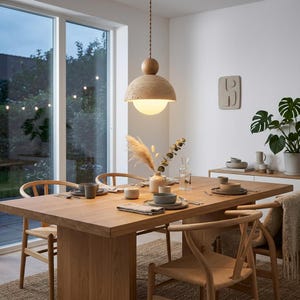 Può includere: Una sala da pranzo con un grande tavolo di legno e sedie. Una lampada a sospensione strutturata è appesa sopra il tavolo. Il tavolo è apparecchiato con piatti, posate e bicchieri. La stanza ha una combinazione di colori neutri.