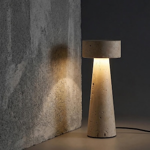Puede incluir: Una lámpara de mesa moderna de hormigón beige, con una parte superior cilíndrica y una base cónica. La lámpara emite una luz cálida y acogedora. La lámpara está conectada a un cable de alimentación negro. La lámpara está junto a una pared gris texturizada.