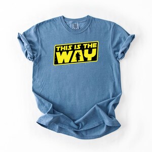 May include: A light blue t-shirt with the text "THIS IS THE WAY" in yellow and black. The design includes a silhouette of a figure. The shirt is made of a soft material.