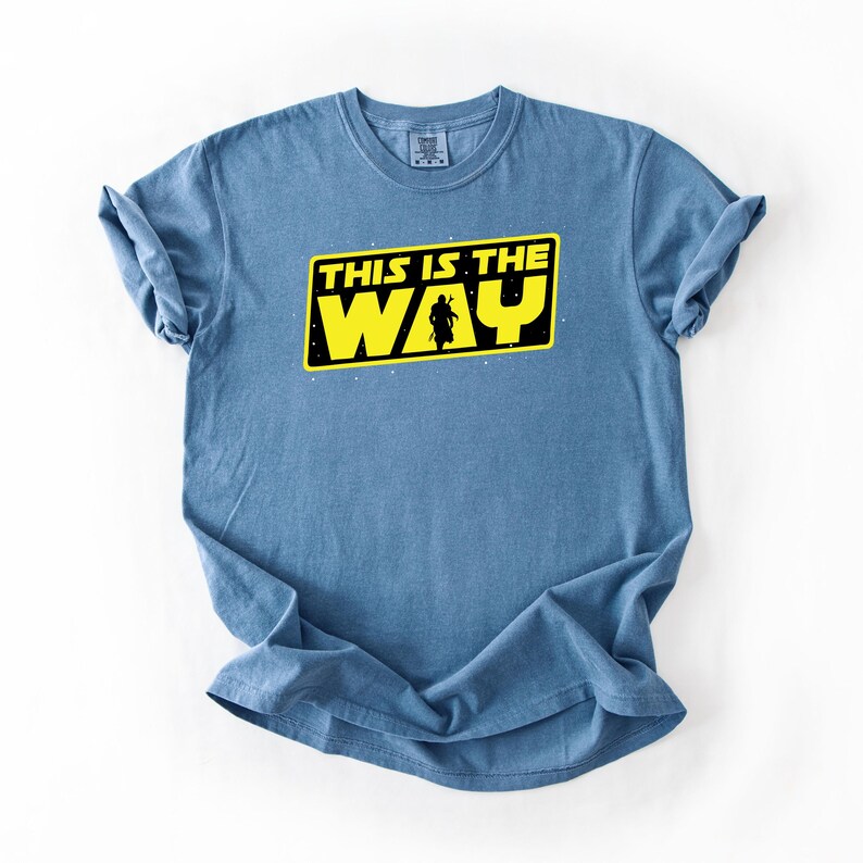 May include: A light blue t-shirt with the text "THIS IS THE WAY" in yellow and black. The design includes a silhouette of a figure. The shirt is made of a soft material.