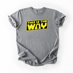 May include: Gray t-shirt with a yellow and black graphic that reads "THIS IS THE WAY". The graphic features a silhouette of a figure. The shirt is made of a soft material and has a crew neck.