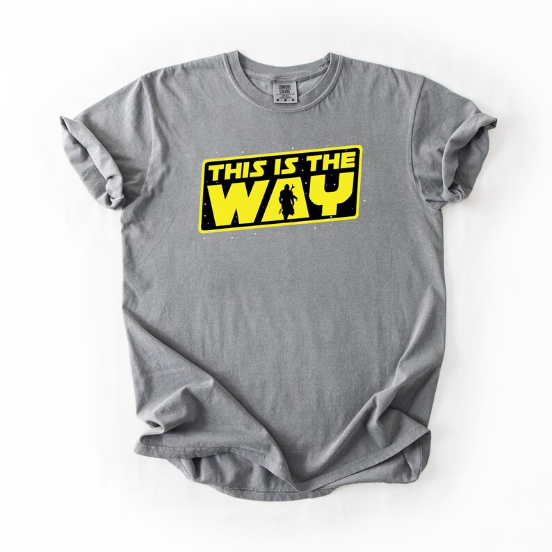 May include: Gray t-shirt with a yellow and black graphic that reads "THIS IS THE WAY". The graphic features a silhouette of a figure. The shirt is made of a soft material and has a crew neck.