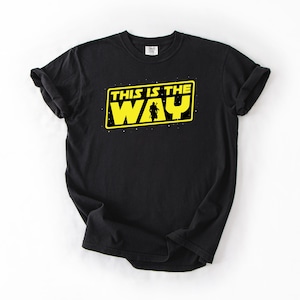 May include: Black t-shirt with a yellow rectangular graphic that reads "THIS IS THE WAY" in white text. A silhouette of a person is in the center of the graphic. The shirt is made of a soft material.