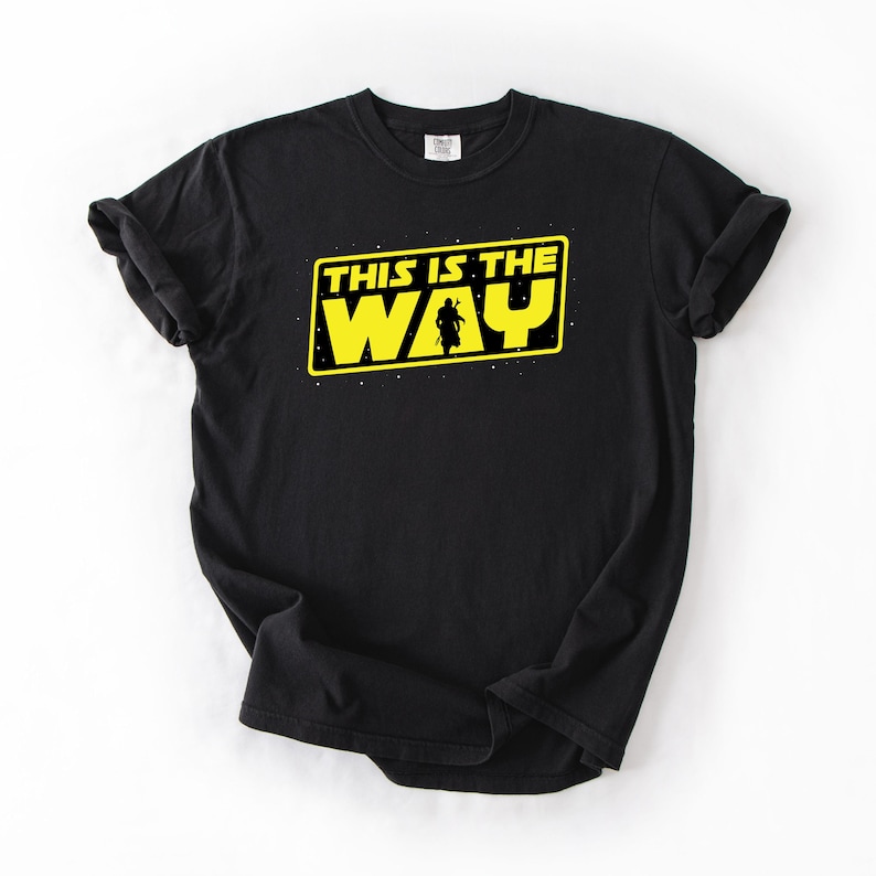 May include: Black t-shirt with a yellow rectangular graphic that reads "THIS IS THE WAY" in white text. A silhouette of a person is in the center of the graphic. The shirt is made of a soft material.