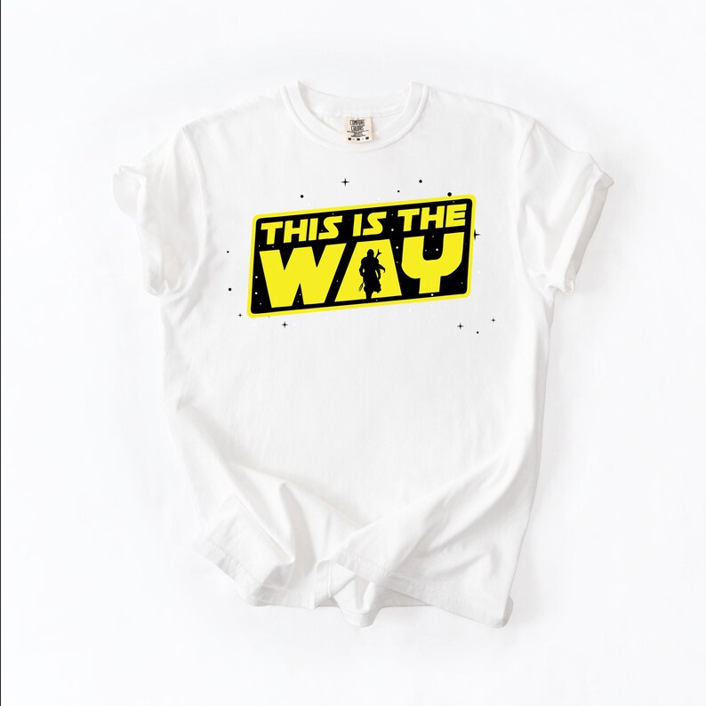 May include: White t-shirt with a yellow and black graphic that reads "THIS IS THE WAY". The graphic features a silhouette of a figure. The shirt is made of a soft material.