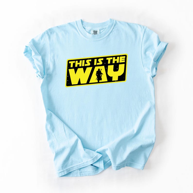 May include: Light blue t-shirt with the text "THIS IS THE WAY" in yellow and black. The graphic includes a silhouette of a figure. The shirt is made of a soft material and has short sleeves.