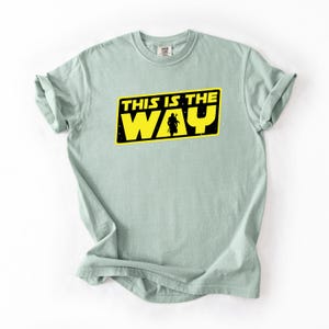 May include: A light green t-shirt with the text "THIS IS THE WAY" in yellow letters inside a black rectangle. A silhouette of a figure is in the center of the rectangle. The t-shirt is folded at the sleeves.