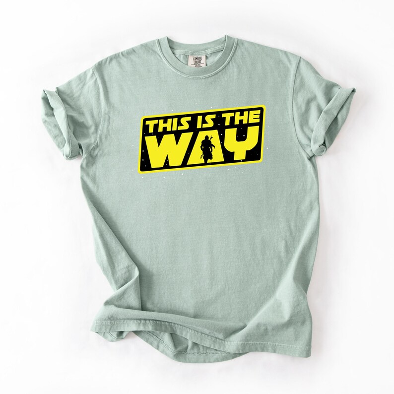 May include: A light green t-shirt with the text "THIS IS THE WAY" in yellow letters inside a black rectangle. A silhouette of a figure is in the center of the rectangle. The t-shirt is folded at the sleeves.