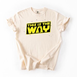 May include: Cream-colored t-shirt featuring the phrase "THIS IS THE WAY" in yellow and black, with a silhouette of a figure. The design is centered on the front of the shirt. The shirt has a crew neck and short sleeves.