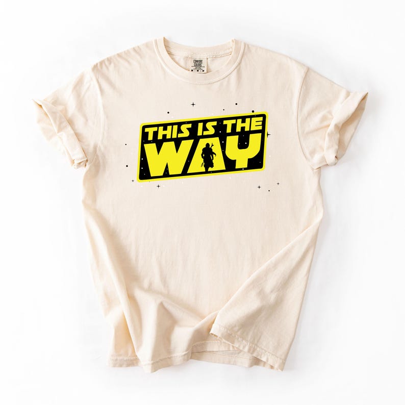 May include: Cream-colored t-shirt featuring the phrase "THIS IS THE WAY" in yellow and black, with a silhouette of a figure. The design is centered on the front of the shirt. The shirt has a crew neck and short sleeves.