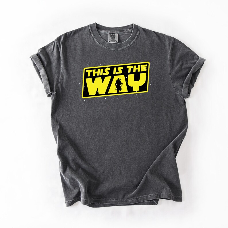 May include: Dark gray t-shirt with the text "THIS IS THE WAY" in yellow and black, featuring a silhouette of a figure. The shirt has a crew neck and short sleeves. The design is centered on the front of the shirt.