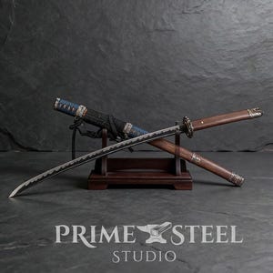 May include: A Japanese katana sword with a curved blade, displayed on a wooden stand. The handle is wrapped in black cord, and the scabbard is blue and black. The text "PRIME STEEL STUDIO" is visible below the sword.