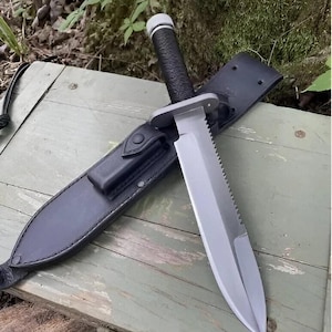 Puede incluir: Un cuchillo grande con una hoja plateada, borde dentado y mango negro, en una funda de cuero negro. El cuchillo tiene una guarda y un mango texturizado. La funda tiene un lazo para el cinturón. El cuchillo está sobre una superficie de madera.
