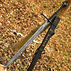 May include: A long, black sword with a patterned blade and a matching sheath. The sword is leaning against a tree branch, with the sheath displaying the name "PINASH" in gold lettering. The blade has a unique, textured design.