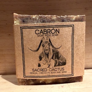Könnte beinhalten: Ein rechteckiges Stück Ziegenmilchseife, eingewickelt in ein beigefarbenes Papieretikett. Das Etikett zeigt eine schwarze Tuschezeichnung einer Ziege sowie den Text CABRÓN GOAT MILK SOAP, SACRED CACTUS, MESCAL, CREOSOTE BUSH AND SAND.