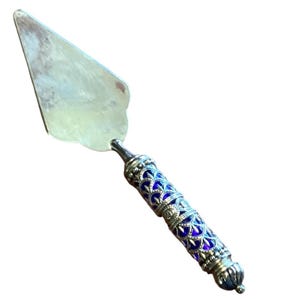 Puede incluir: Una espátula plateada para pasteles con un mango decorativo. El mango presenta un diseño azul y plateado. La espátula tiene una hoja triangular y un mango detallado y ornamentado. La espátula está diseñada para servir pasteles y repostería.