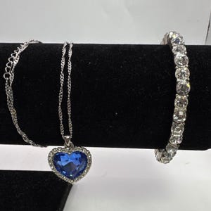 Puede incluir: Un collar plateado con un colgante azul en forma de corazón rodeado de piedras transparentes. También se muestra una pulsera a juego con piedras transparentes. Ambas piezas están sobre una superficie de terciopelo negro.