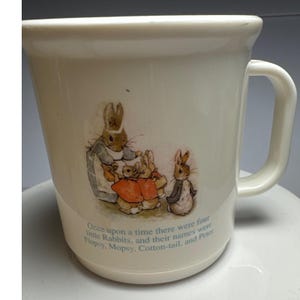 May include: A cream-colored mug featuring a printed illustration of four rabbits from the Peter Rabbit story. Below the illustration, the text reads: Once upon a time there were four little Rabbits, and their names were Flopsy, Mopsy, Cotton-tail, and Peter.