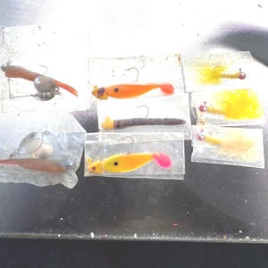 Op de afbeelding: Een verzameling van acht viskunstaasjes in individuele doorzichtige plastic hoesjes. De set bevat zacht plastic kunstaas in oranje, geel en bruin, sommige met metalen spinnerbladen, en diverse gele gevederde jigs met rode koppen en haken.