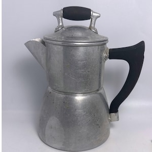 Pode incluir: Uma cafeteira de alumínio escovado com uma base larga e cónica, um bico vertedor proeminente e uma tampa abobadada. A cafeteira inclui uma pega lateral preta resistente ao calor e um pequeno botão preto no topo da tampa para a levantar.