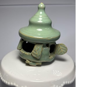 Puede incluir: Un quemador de incienso de cerámica esmaltada en verde claro con forma de pequeña pagoda o casa. La estructura presenta un techo puntiagudo, ventanas arqueadas abiertas y detalles decorativos en forma de hoja en la base con bordes de arcilla clara.
