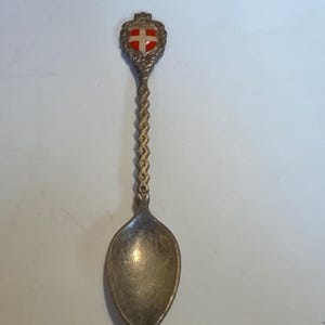 May include: A silver-colored souvenir spoon with a decorative handle and a small shield-shaped emblem at the top. The emblem features a red cross on a white background. The spoon's handle has a twisted rope design.