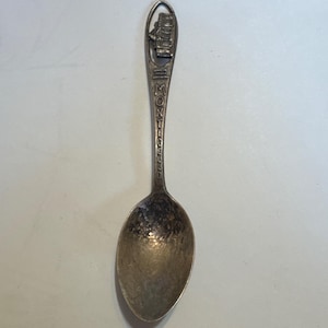 May include: A silver spoon with a detailed design at the handle's top, featuring a building and the word "MONTICELLO". The spoon's handle is slender, leading to a rounded bowl. The spoon is likely a souvenir or collectible item.