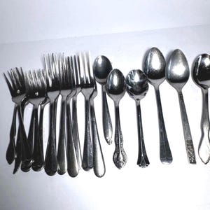 Peut inclure: Une collection de couverts en acier inoxydable comprenant plusieurs fourchettes et cuillères de tailles et de designs variés. L'ensemble présente des surfaces métalliques polies avec des motifs en relief sur certains manches et cuillerons.