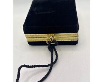 Könnte beinhalten: Eine rechteckige Schmuckschatulle aus schwarzem Samt mit einem goldfarbenen Metallrand entlang der mittleren Naht. Die Schatulle verfügt über eine kleine goldfarbene Metallöse an der Vorderseite, an der ein schwarzer Kordelgriff befestigt ist.