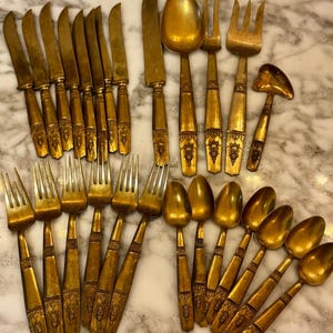 May include: An assortment of antique gold-colored flatware, including knives, forks, spoons, and a serving utensil. The handles feature ornate detailing. The arrangement is on a marble surface.