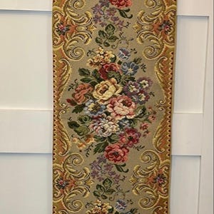 May include: A rectangular tapestry with a floral design. The tapestry features a beige background with a repeating pattern of colorful flowers in shades of pink, red, yellow, and blue. The edges are trimmed with a gold border and a tassel.