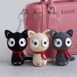 May include: Three crocheted cat keychains in black, beige, and dark gray. Each cat features large white circular eyes with black pupils, embroidered eyelashes, a red crocheted scarf around the neck, and small white embroidered paws at the base.