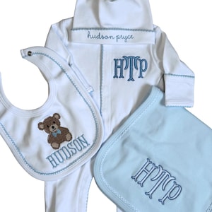 Könnte beinhalten: Ein weißes Baby-Set bestehend aus einem langärmeligen Strampler mit passender Mütze, einem Lätzchen und einem Spucktuch. Strampler und Mütze sind mit dem Namen hudson pryce und dem Monogramm HTP in Blau bestickt. Das Lätzchen zeigt einen braunen Bären.