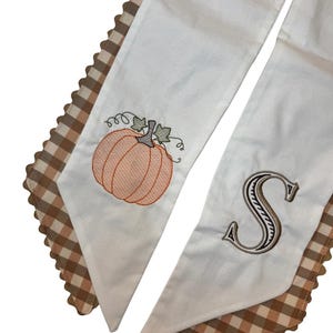 May include: Two white fabric strips with brown and tan gingham borders featuring scalloped edges. One strip displays an embroidered orange pumpkin with green leaves, while the other strip features a large, embroidered letter S in brown.