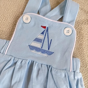 May include: Light blue baby romper with a sailboat design. The romper features a sailboat embroidered on the chest with white and blue stripes, a red flag, and white trim. It has two white buttons on the straps.