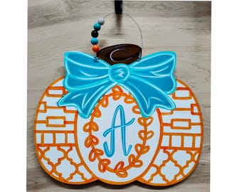 May include: A wooden pumpkin-shaped door hanger with a turquoise bow and the letter "A" in a wreath. The pumpkin is white with orange accents and a decorative pattern. A beaded handle and a brown top complete the design.