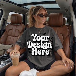 Op de afbeelding: Donkergrijs t-shirt met de tekst "Your Design Here" in een retro lettertype. Het shirt wordt gedragen door een persoon die in een auto zit. De persoon draagt een zonnebril en ringen. Een drankje in een doorzichtige beker wordt in de hand gehouden.