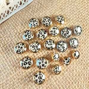 May include: A collection of round buttons featuring a cream-colored base with a black and brown leopard print pattern. The buttons have smooth, glossy surfaces and are framed by metallic gold or silver rims, arranged in a scattered cluster.
