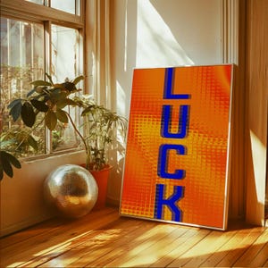 May include: A framed art print displaying the word "LUCK" in blue block letters on an orange, textured backdrop. The print is propped against a wall in a room with a disco ball, plants, and a window.
