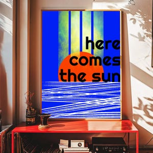 May include: A framed art print with a colourful design. The artwork features a bright orange sun, blue water, and the words "here comes the sun" in black. Vertical blue and yellow stripes are in the background. The print is displayed on a red table.