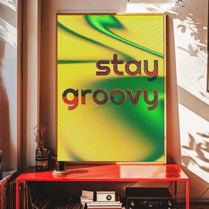 May include: A framed art print with the words "stay groovy" in a retro font. The artwork features a yellow and green abstract background. The frame is gold, and the print is displayed on a red cabinet.
