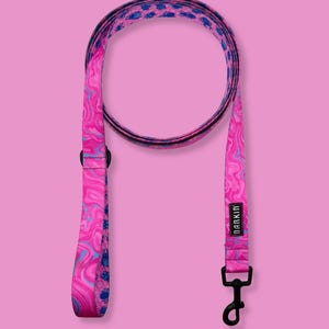 May include: A pink and blue dog leash featuring a swirling marble pattern on one side and a repeating blue beetle insect print on the other. The leash includes a black metal swivel snap hook and a black rectangular label with the text BARKIN.
