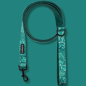 May include: A teal dog leash featuring a repeating pattern of green leaves and ice cubes. The leash includes a black metal swivel snap hook, a black D-ring, and a rectangular black fabric tag with the white text BARKIN printed on it.