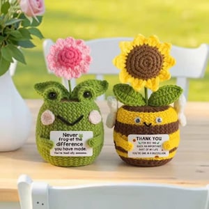 May include: Two handmade crochet planters: a green frog with a pink flower and a yellow and brown bee with a sunflower. Each holds a small sign with a message. The frog's sign reads "Never frog-et the difference you have made."