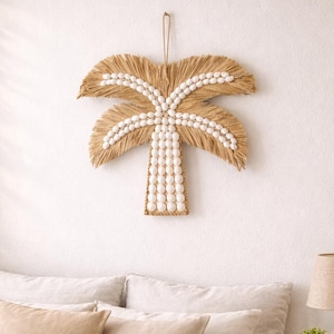 Könnte beinhalten: Eine Wanddekoration in Form einer Palme aus Naturfasern und weißen Perlen. Die Palme hängt über einem Bett mit beiger Bettwäsche und Kissen. Ein Nachttisch aus Holz mit einer Lampe und einer kleinen Pflanze befindet sich neben dem Bett.