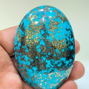 May include: An oval-shaped polished turquoise gemstone featuring a vibrant sky-blue base colour with intricate, metallic gold and dark brown matrix patterns distributed across the surface, creating a unique contrast of textures and colours.
