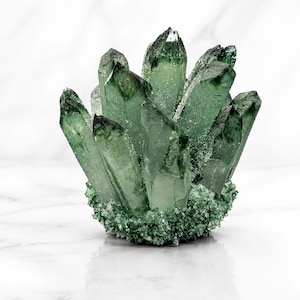 Pode incluir: Um aglomerado de cristais de quartzo verde-escuro translúcidos que se erguem de uma base de formações minerais verdes menores e texturizadas. As estruturas cristalinas prismáticas e pontiagudas variam em altura e tom, exibindo uma textura de superfície natural, irregular e cristalina em todo o espécime.