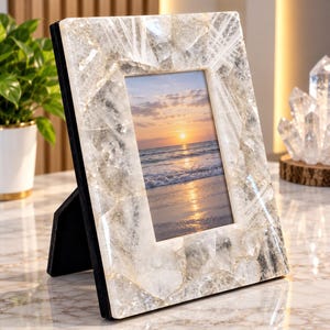 May include: A rectangular picture frame featuring a border constructed from numerous clear, jagged quartz crystal pieces. The frame has a black backing and stand, and it holds a photograph of a sunset over the ocean with waves hitting the sand.