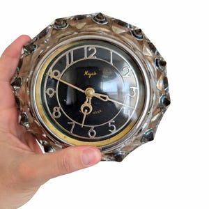 May include: A vintage round clock with a black face and gold hands and numerals. The clock face has the word "Majak" and "USSR" printed on it. The clock is encased in a clear, faceted glass frame.