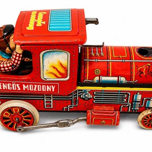 Könnte beinhalten: Ein roter Vintage-Blechspielzeugzug mit der Aufschrift "Csengős Mozdony" an der Seite. Der Zug hat eine Fahrerfigur, ein detailliertes Motordesign und ein Büffel-Emblem. Die Räder sind rot und weiß.